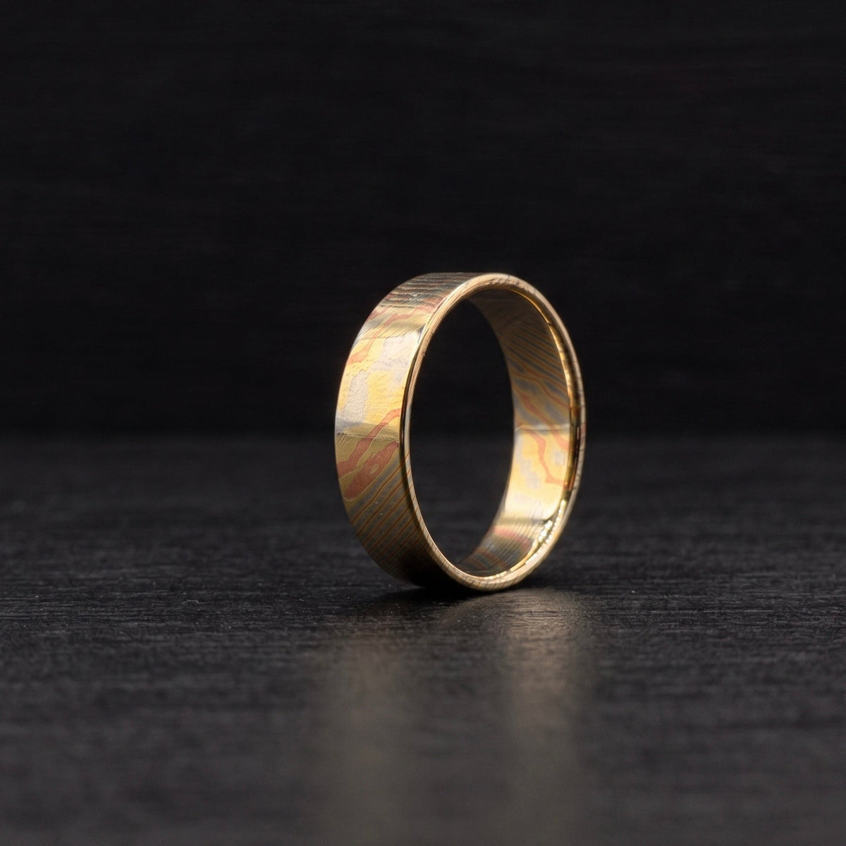 A single gold-toned ring with a textured, hammered finish and a subtle wavy pattern inside, standing upright on a dark, textured surface against a dark background.