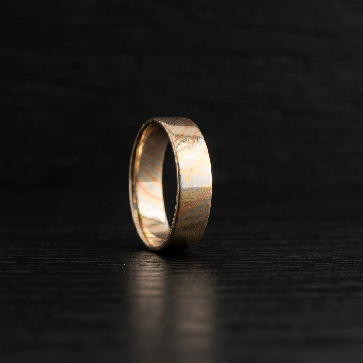 A single gold band ring with a hammered or textured finish, standing upright on a dark, textured surface.